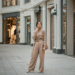 A stylish woman wearing a neutral-toned outfit, posing thoughtfully in front of an Aritzia store.