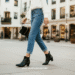 A stylish woman wearing black kitten heel ankle boots with cropped blue jeans and a white blouse, walking down a city street.