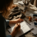 An aspiring fashion designer sketching a dress design in a sunlit studio with fabric swatches and tools on the table.