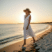 A stylish woman in a white linen dress and straw hat walking along a beach in the Hamptons at sunset.