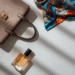 A stylish flat lay of iconic accessories from several top luxury fashion brands, including a handbag, a silk scarf, and a bottle of perfume.