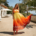 A woman wearing a beautiful floor-length orange ombre dress, with colors blending from dark orange at the bottom to light yellow at the top, standing in a sunny outdoor setting.