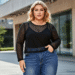 A confident plus-size woman smiling while wearing a stylish black long-sleeved mesh top layered over a camisole, paired with blue jeans.