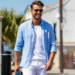 A stylish man wearing a light blue linen shirt and white chino shorts, smiling confidently on a sunny day.