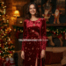 A smiling woman wearing a festive red velvet Christmas dress, standing next to a decorated Christmas tree.
