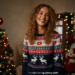 A person smiling while wearing a festive red and green Christmas sweater decorated with reindeer and snowflakes.