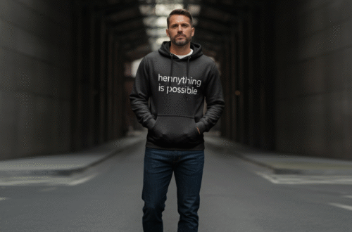 A person wearing a stylish black 'hennything is possible hoodie' paired with jeans, standing against a neutral urban background.